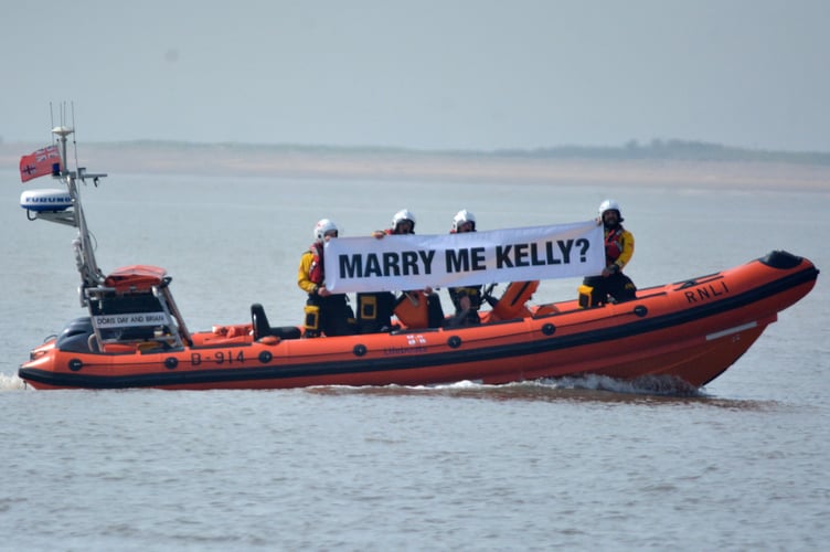 Burnham's Atlantic 85 displaying Lyndon's message to Kelly. Photo released May 15 2024. RNLI volunteer surprises partner with unique proposal - with lifeboat crew holding up huge banner on the waves.Lyndon and Kelly were walking along Burnham-On-Sea beach on Sunday (12 May) - when a lifeboat appeared on the waves with RNLI crew holding a three metre banner stating: Marry me, Kelly?.After nearly 20 years together, Lyndon, the Burnham-on-Sea RNLI Deputy Press Officer, decided there was no better place to get down on one knee than on the coastline in front of RNLI's Lower Lighthouse.Lyndon, who had spent spent weeks planning the very special moment with the help of crew, said: "Thankfully she said yes!"